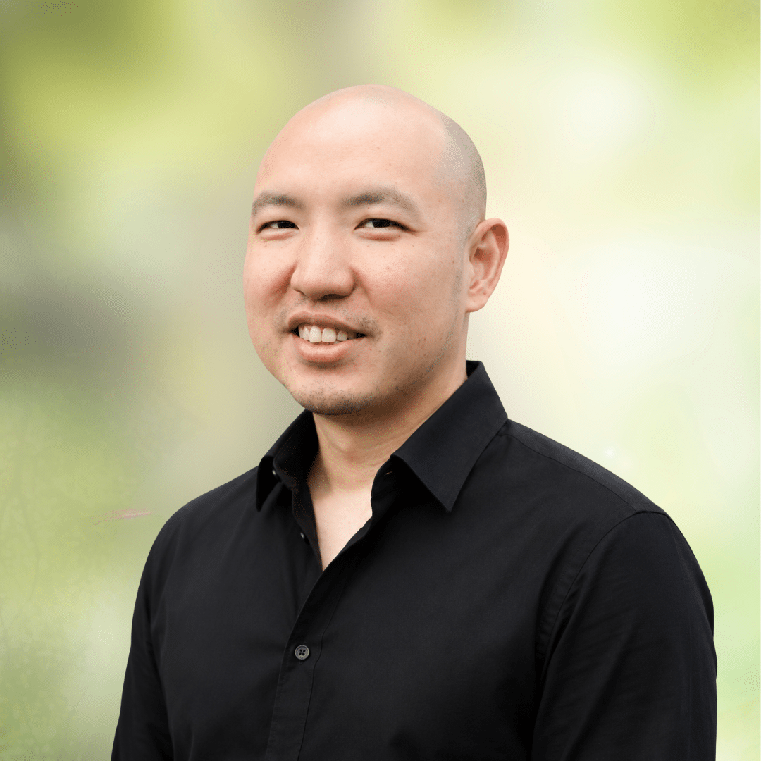 Smiling bald man in a black button-down shirt, portrait outdoors with a soft green background.
