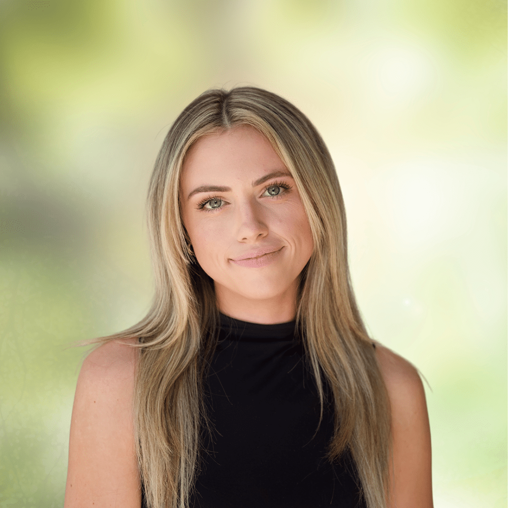 Portrait of a young woman with long blonde hair in a black sleeveless top, smiling softly outdoors, with a green blurred background.
