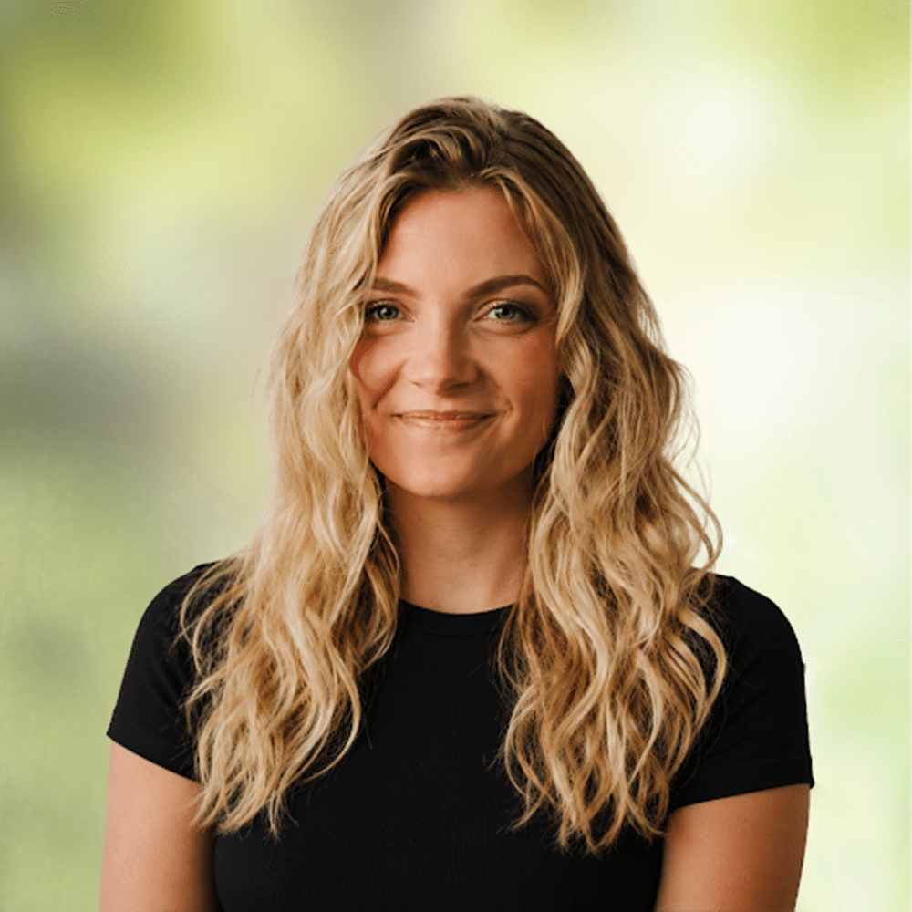 Portrait of a smiling woman with long wavy blonde hair wearing a black shirt against a soft green background.